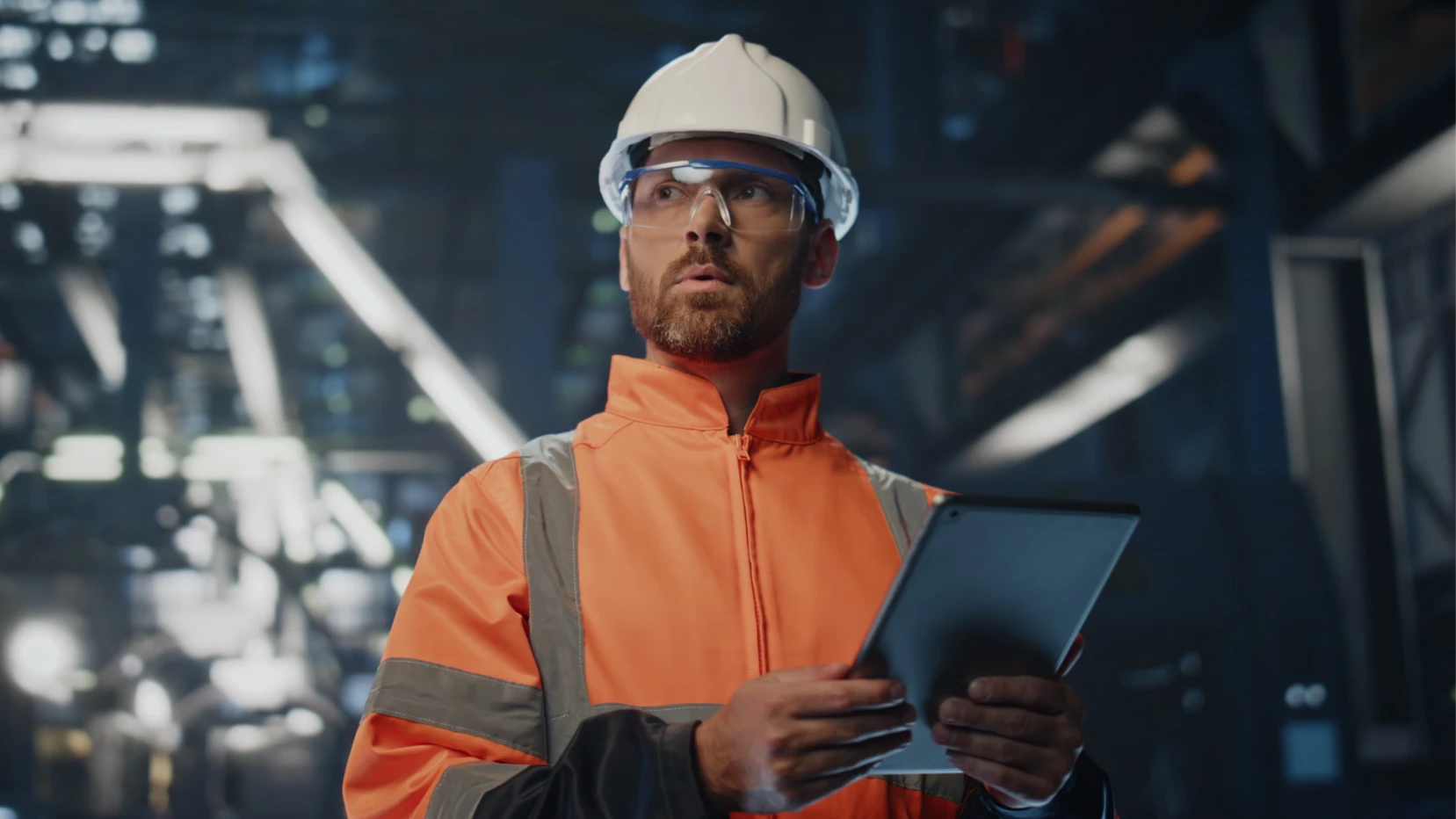 Industrial technician in an orange jacket and white hard hat holding a tablet inside a plant